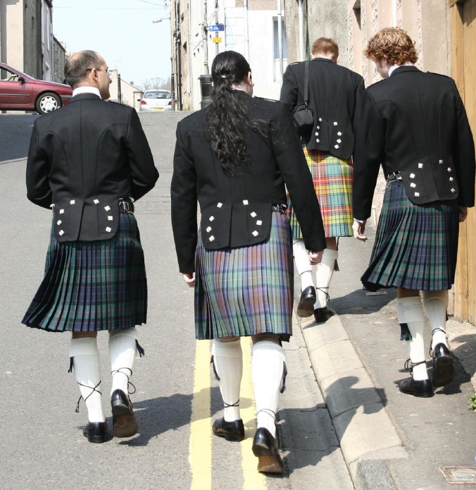 Four men wearing kilts walk down a city street.