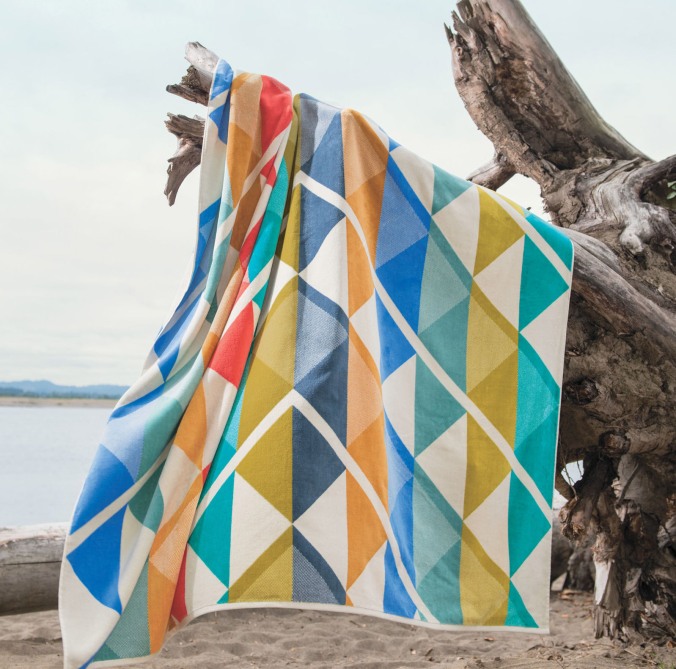 A Pendleton geometric spa towel hangs on a piece of driftwood on the seashore.