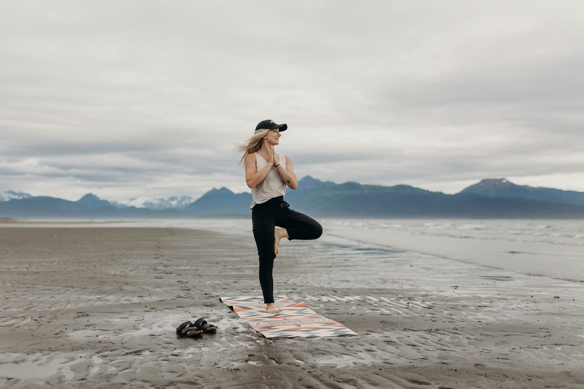 A young woman does a pose on a Pendleton x Yeti yoga mat, on a beach in front of a distant mountain range. 