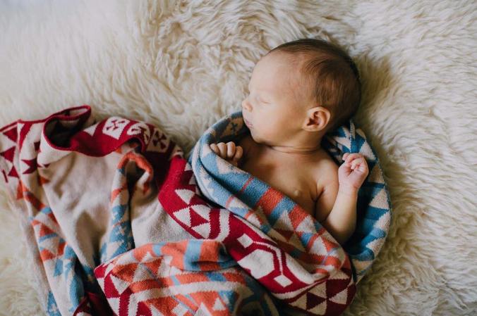 A newborn baby, wrapped in a knitte pendleton baby blanket, lies on a soft white sheepskin.