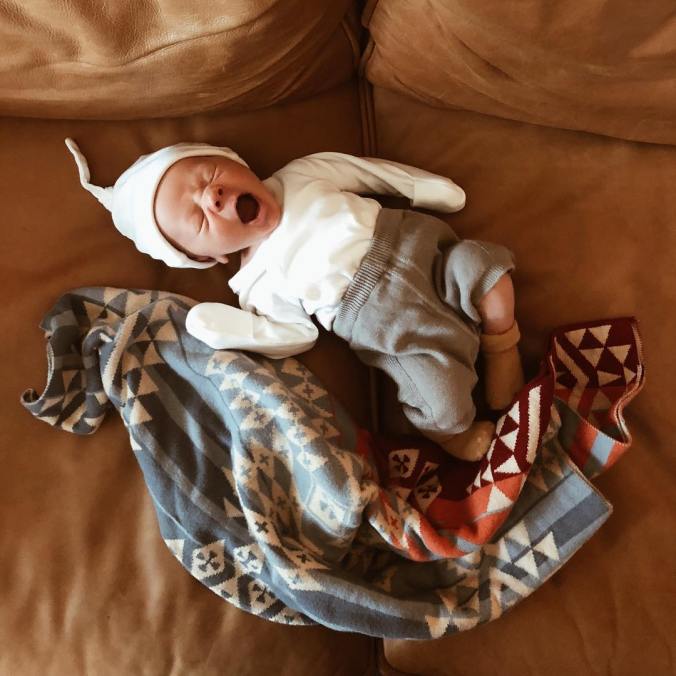A newborn baby lies on a leather sofa next to a knitted pendleton baby blanket. The baby is wearing soft knitted clothing and a stocking cap, and the baby is yawning. 