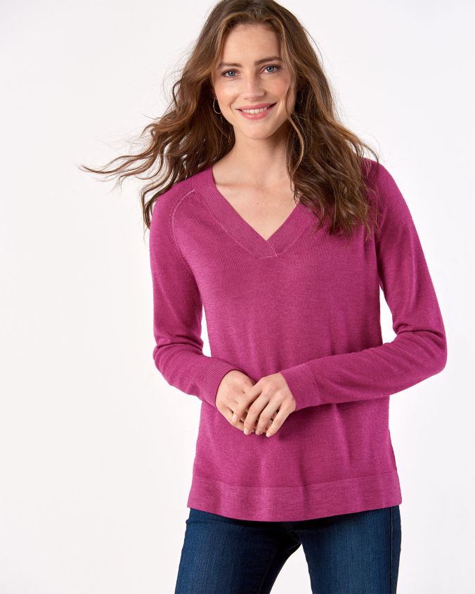 A woman with long brown hair and blue eyes stands before a white background, her hands held in front of her stomach. She is wearing a plum colored sweater and jeans.