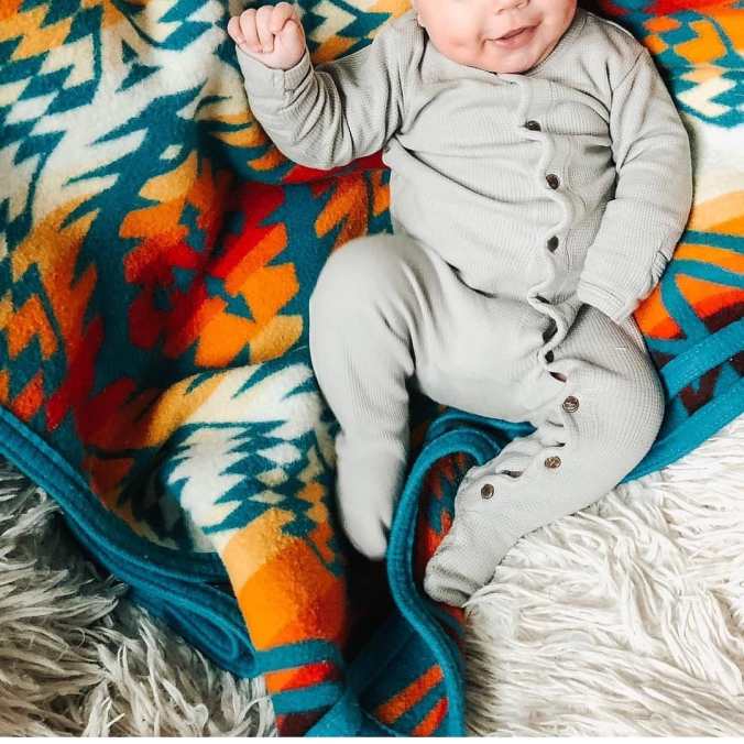 A smiling baby lies on a Pendleton Thunder and Lightning blanket. The baby is wearing a light grey thermal knit onesie with small wooden buttons.