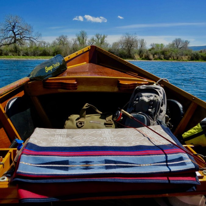 Photo taken from within a hand-built wooden drift boat, showing a folded Pendleton Bighorn blanket on the seat, and gear packed below the boat's prow, which is poised above the waters of Montana's Bighorn river.