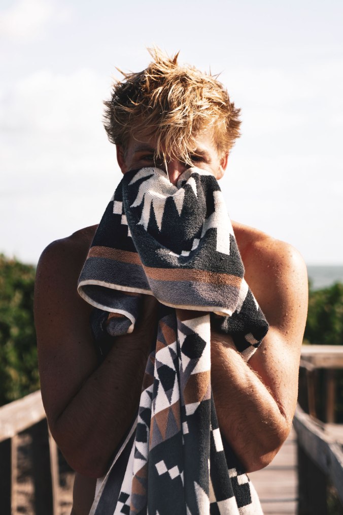 A young man with brushy blond hair dries his face with a Pendleton Spider Rock patterned beach towel. 